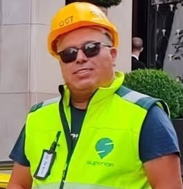 Man wearing sunglasses, a yellow safety helmet, and a bright yellow reflective vest with a logo, standing outdoors.