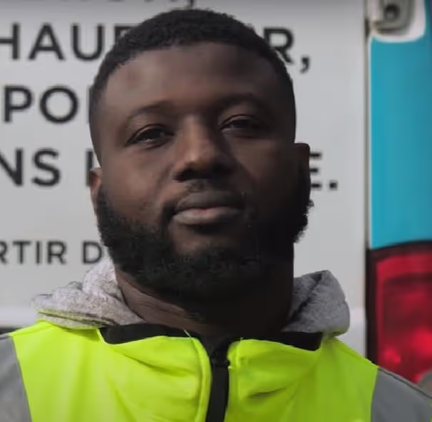 Portrait of a man with a beard wearing a neon yellow safety vest and hoodie, standing outdoors with a vehicle in the background.