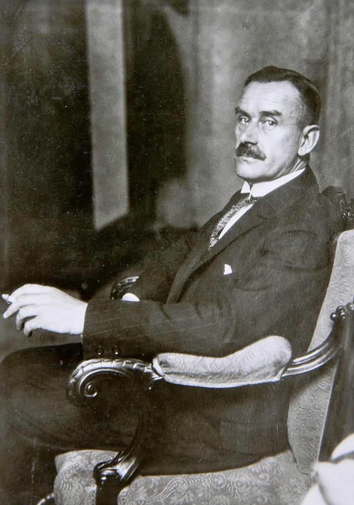 Thomas Mann in a formal suit sits elegantly in a vintage armchair, holding a cigarette.