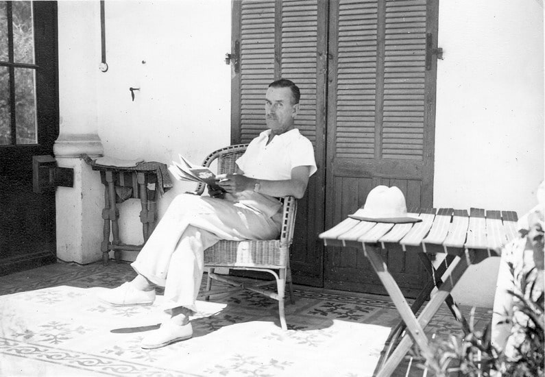 Thomas Mann sits relaxed in a wicker chair, reading a book on a porch with wooden shutters and a small table nearby. A hat rests on the table.