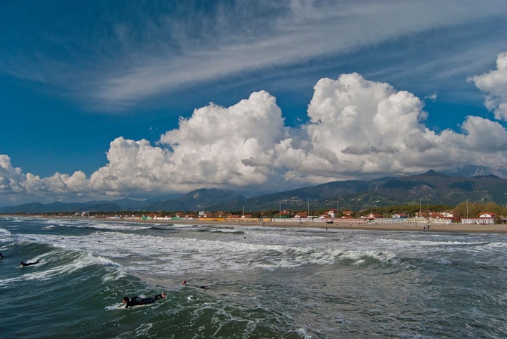 A scenic beach view with surfers, rolling waves, colorful houses, and dramatic clouds against a mountain backdrop.