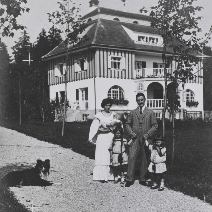 A vintage family portrait outside a large, two-story house, featuring two adults, three children, and a dog on a gravel path.
