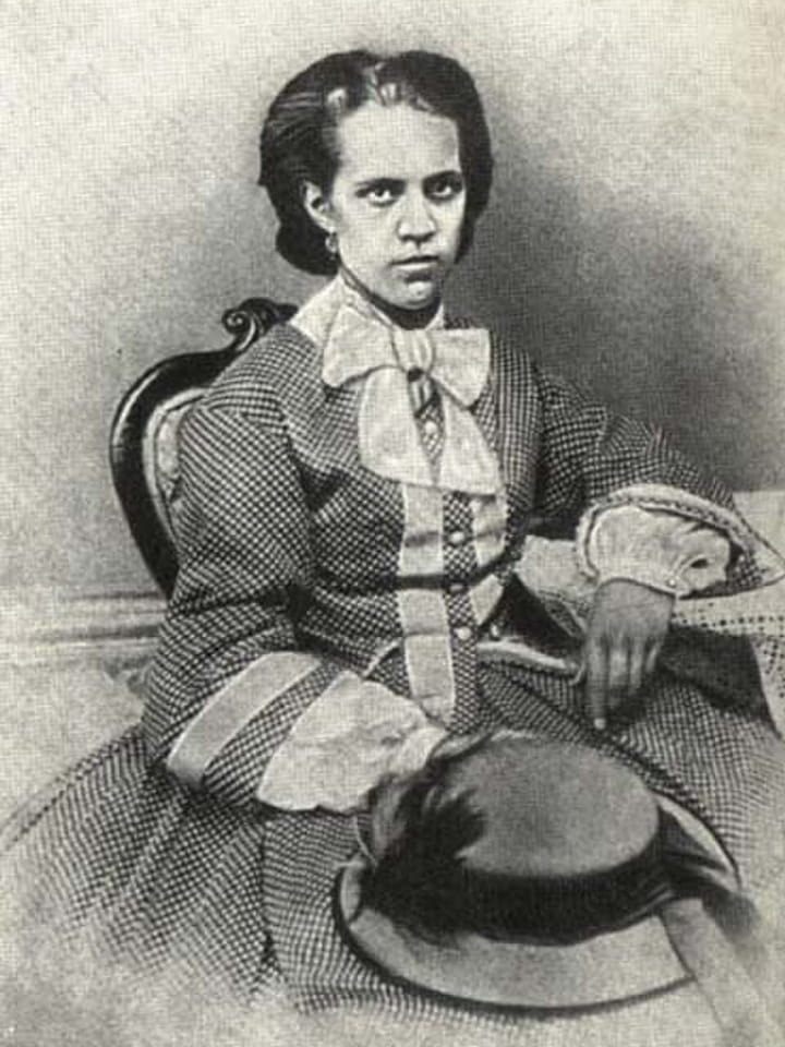 Young woman with dark hair in a patterned dress sitting on a chair holding a hat