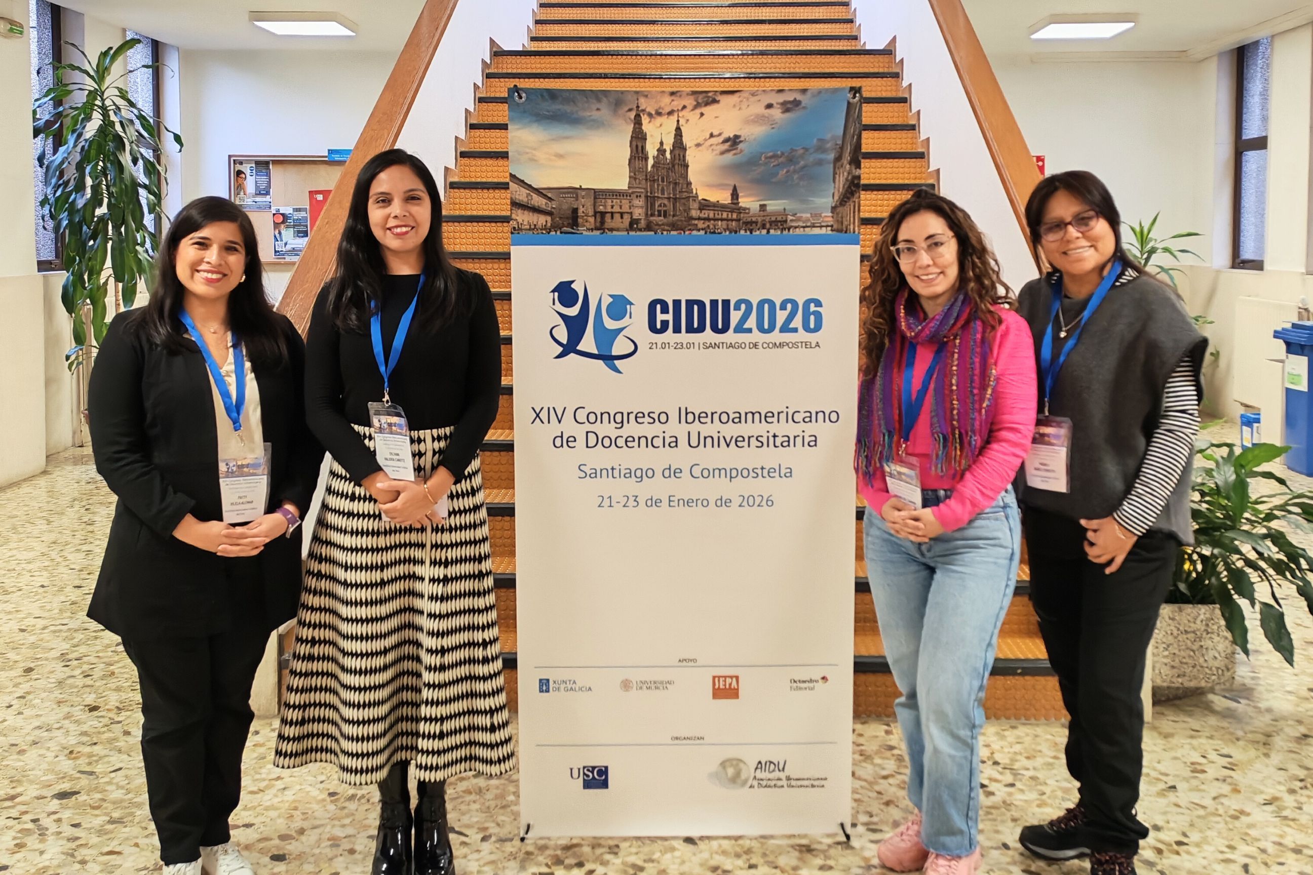 Grupo de cuatro representantes de la Dirección de Asuntos Estudiantiles (DAES) de la PUCP posan frente a un panel del CIDU 2026, ubicado al pie de una escalera en un espacio interior. Todas llevan credenciales del evento y sonríen a la cámara. En el panel se lee “XIV Congreso Iberoamericano de Docencia Universitaria – Santiago de Compostela, 21-23 de enero de 2026”, acompañado de una imagen de la catedral de la ciudad.