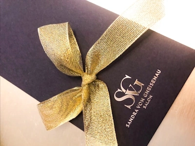 Schwarzes Geschenkumschlag mit goldenem Band und dem Schriftzug Sandra von Gneisenau Salon.