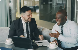 Two businessmen in suits sitting at a table with a laptop and coffee cups, smiling and having a conversation.
