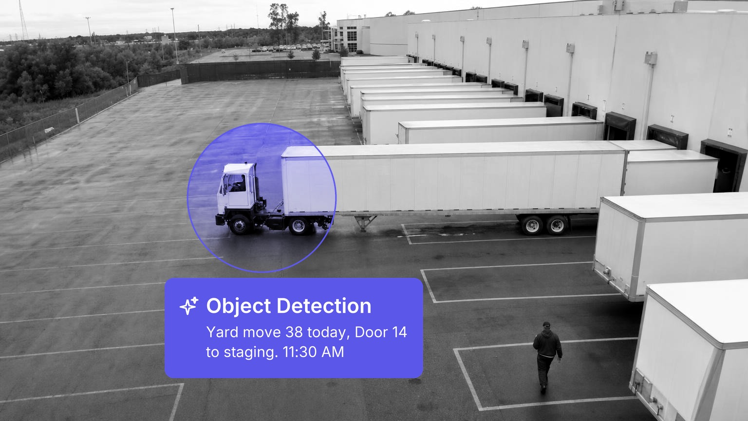 A yard truck pulling a plain white trailer across the wet asphalt yard of a distribution center past a row of trailers backed into dock doors, a worker walking away between the parking stripes, in black and white with a Spot AI detection overlay.
