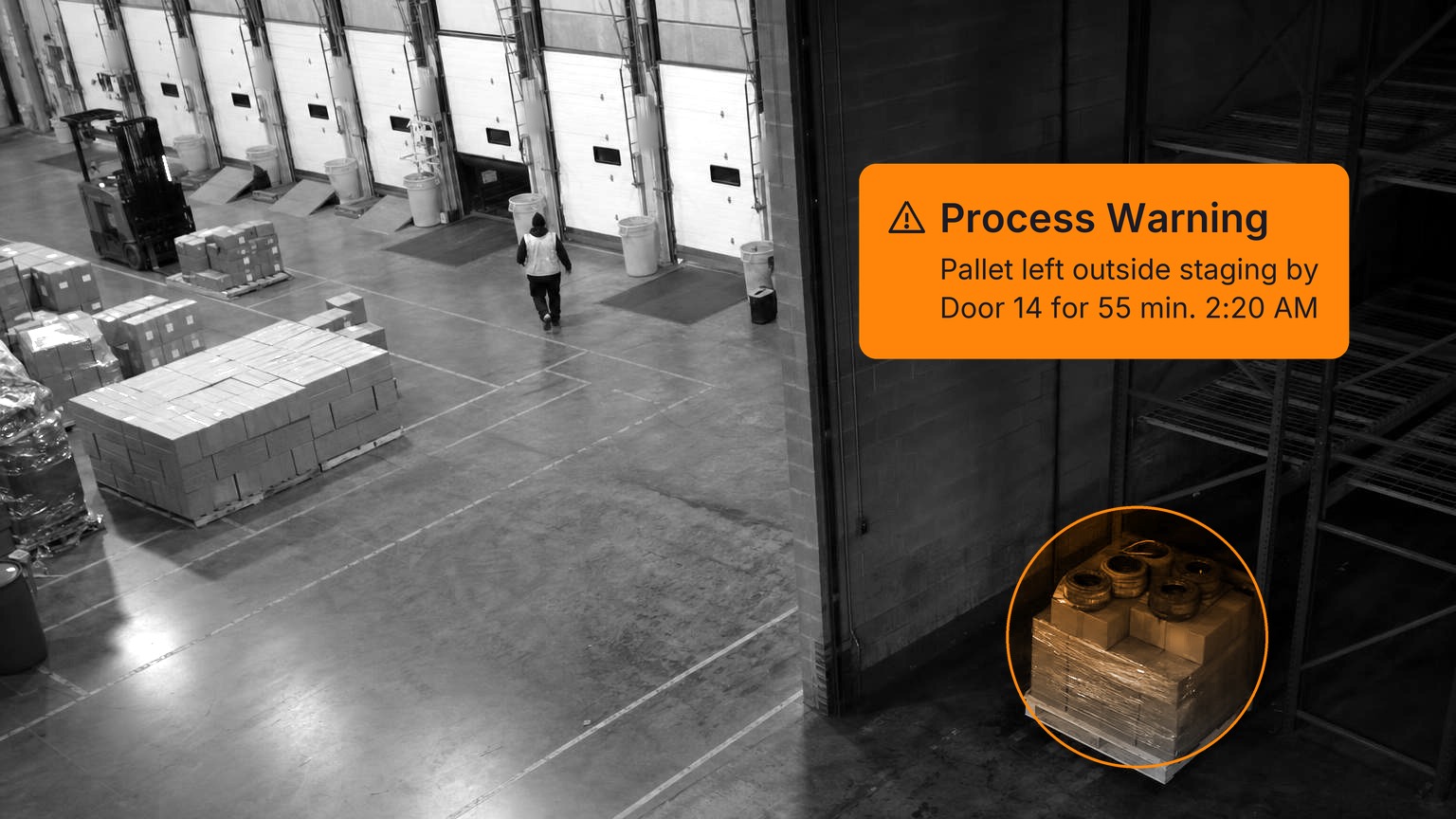A shrink-wrapped pallet of cartons and brake drums sits alone in a dim corner beside a rack, away from the staging lanes, while a worker in a hi-vis vest, seen from behind, walks toward the dock doors of a distribution center, in black and white with a Spot AI detection overlay.