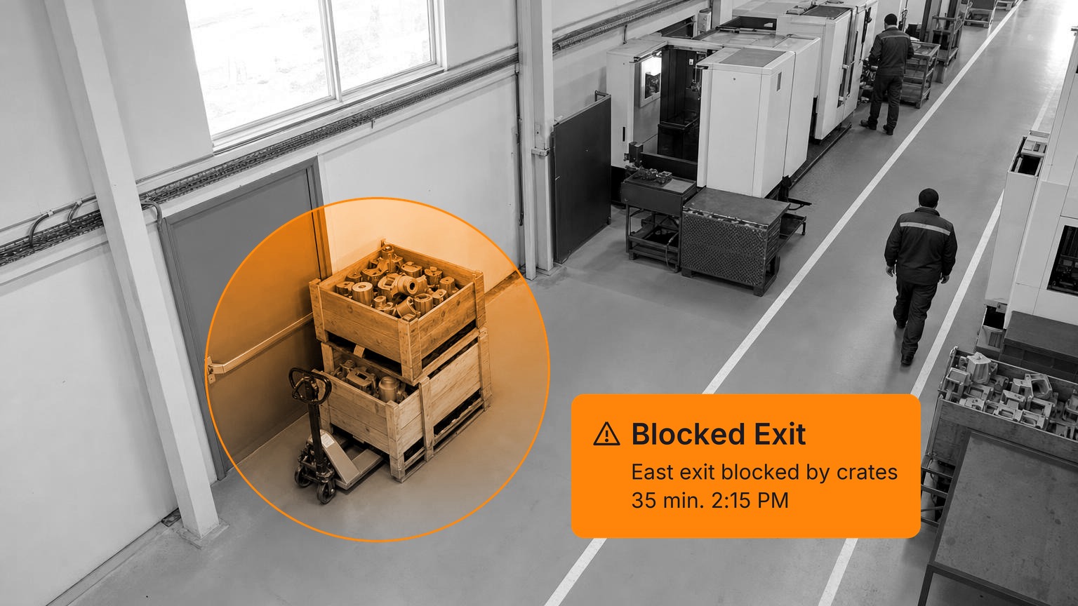 Two stacked wooden crates of metal castings and a pallet jack left against a grey emergency exit door in a machining hall, with operators walking away toward the CNC machines, in black and white with a Spot AI detection overlay.
