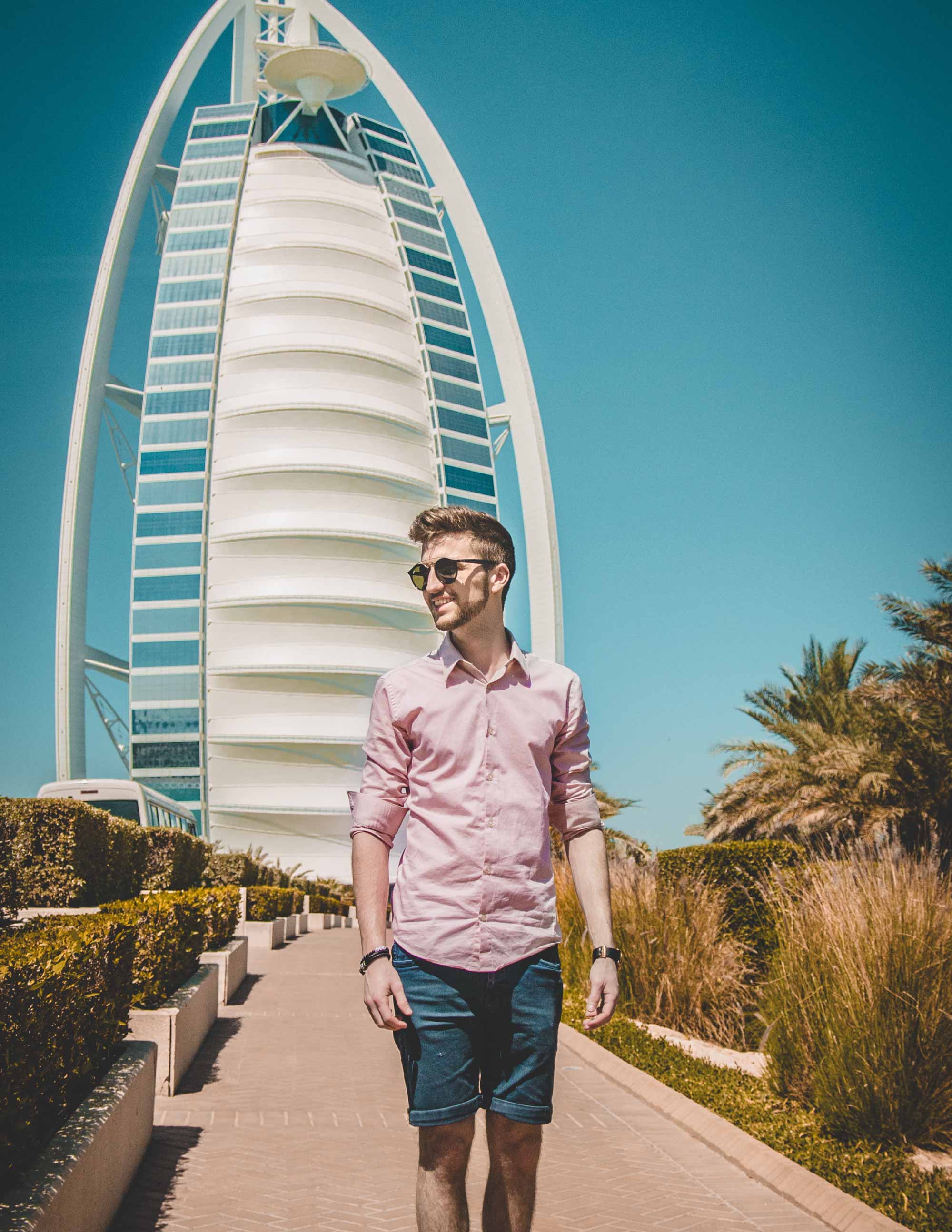 a young man in a pink shirt walks along a planted path in front of the Burj Al Arab in Dubai