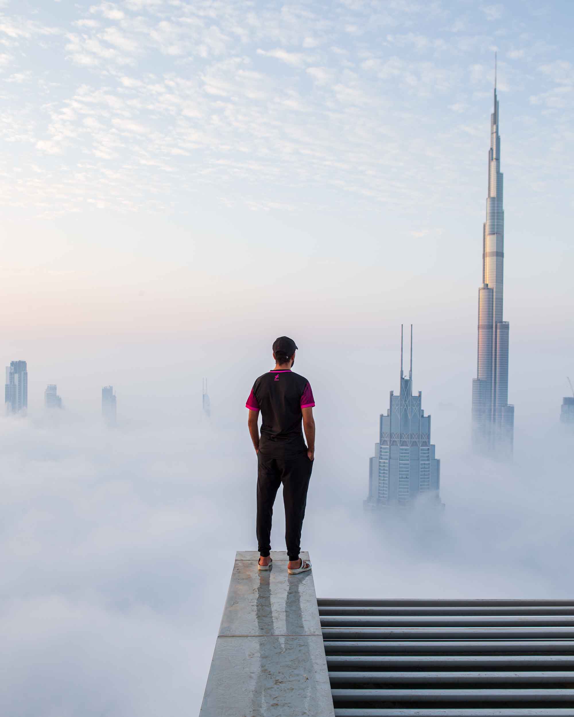 A man stands on top of an extremely high building, overlooking the skyscrapers of Dubai push their way through a thick white cloud.