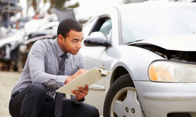 Insurance adjuster examining a damaged car and taking notes on a clipboard at an accident scene.