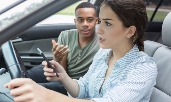 Passenger gesturing with concern as the driver looks at a phone while driving, illustrating distracted driving and passenger safety concerns.