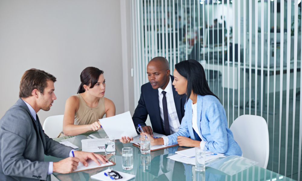 Group of four professionals in business attire sitting around a glass table in a modern office, reviewing documents and discussing legal or business matters.