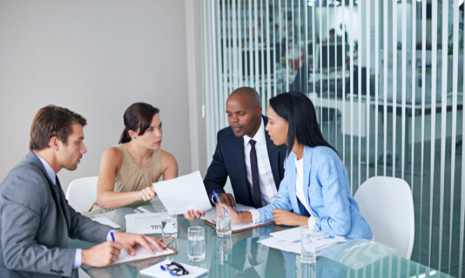 Group of four professionals in business attire sitting around a glass table in a modern office, reviewing documents and discussing legal or business matters.