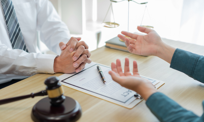 Close-up of legal scales and a gavel on a desk with two people in business attire shaking hands in the background, symbolizing a legal agreement or settlement.