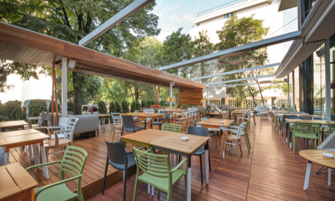 Outdoor restaurant patio with wooden flooring and various dining tables and chairs arranged under a partially covered roof, surrounded by trees and greenery.
