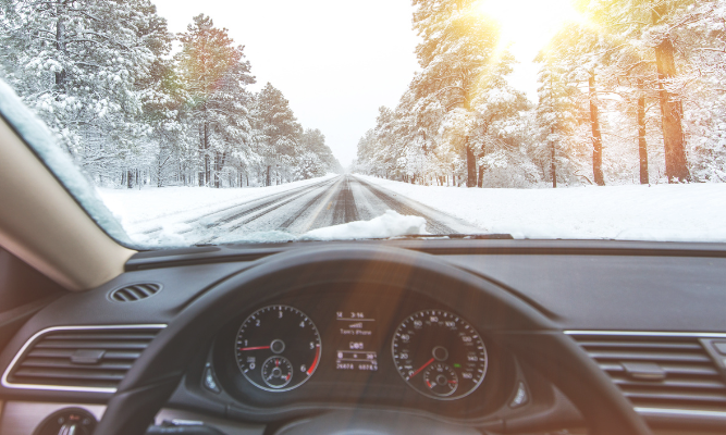 View from inside a car driving down a snow-covered road surrounded by trees with bright sunlight shining through, representing winter driving conditions.