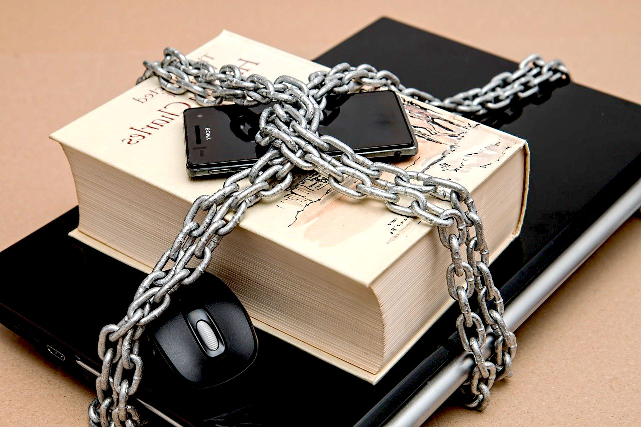 Imagem de dispositivos eletrônicos, celular, notebook e um livro acorrentados