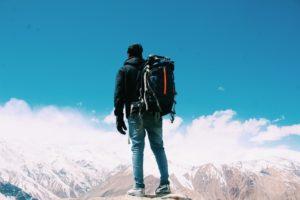 Na foto você pode ver um homem de pé em uma montanha. Ao fundo estão os picos de outras montanhas, nuvens brancas e o céu azul. O homem carrega uma mochila nas costas.
