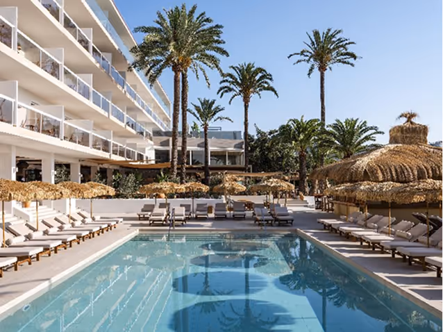 Outdoor hotel pool area with lounge chairs, thatched umbrellas, and tall palm trees under a clear blue sky.