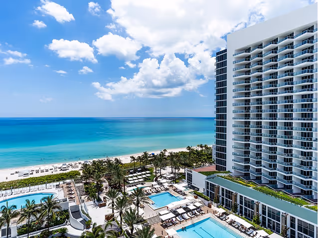 Oceanfront resort with swimming pools, palm trees, and a tall white hotel building under a partly cloudy sky.