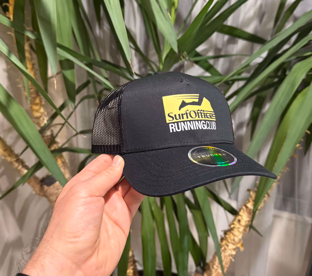 Mano che regge un cappello da camionista nero con il logo giallo e bianco del Surf Office Running Club davanti a piante verdi e frondose.