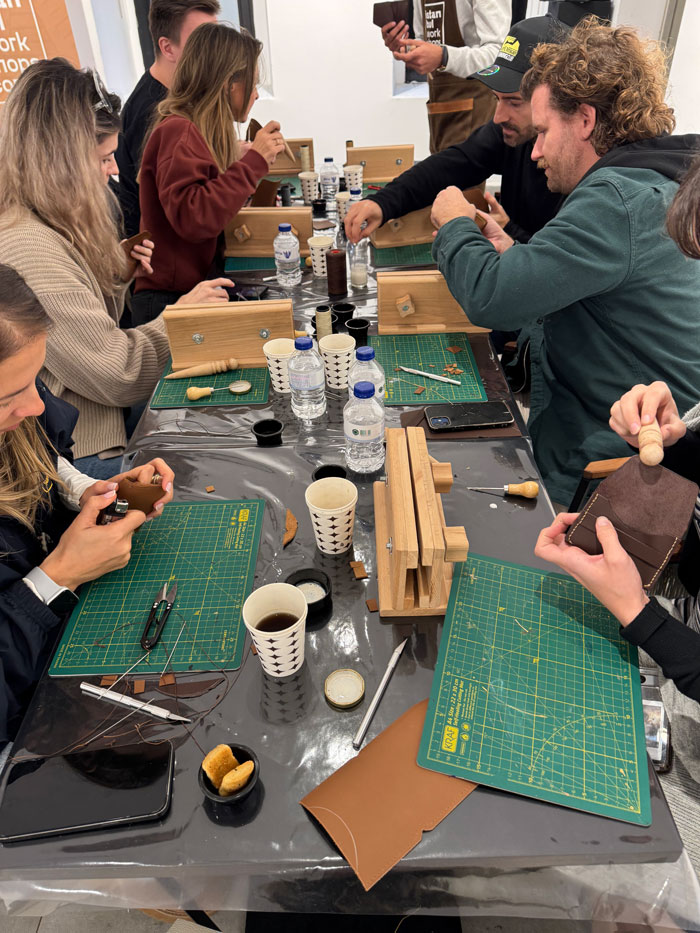 Un gruppo di persone sedute attorno a un tavolo realizzano oggetti in pelle utilizzando utensili, tappetini da taglio e supporti in legno.