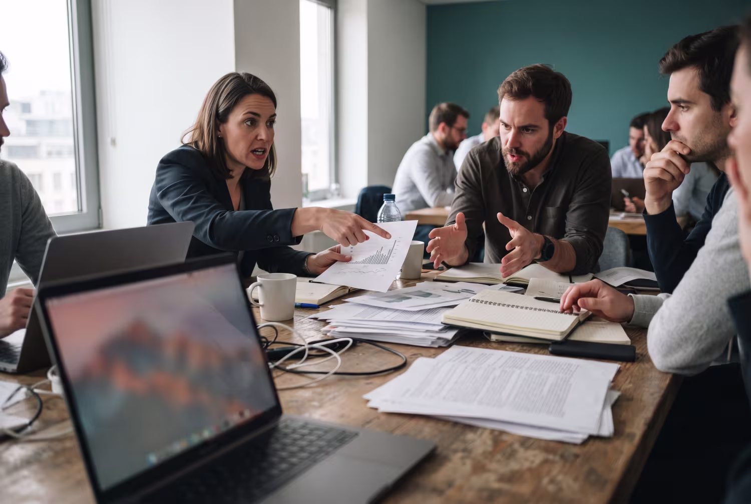 Professionals discussing around cluttered table with open laptop showing colorful abstract display