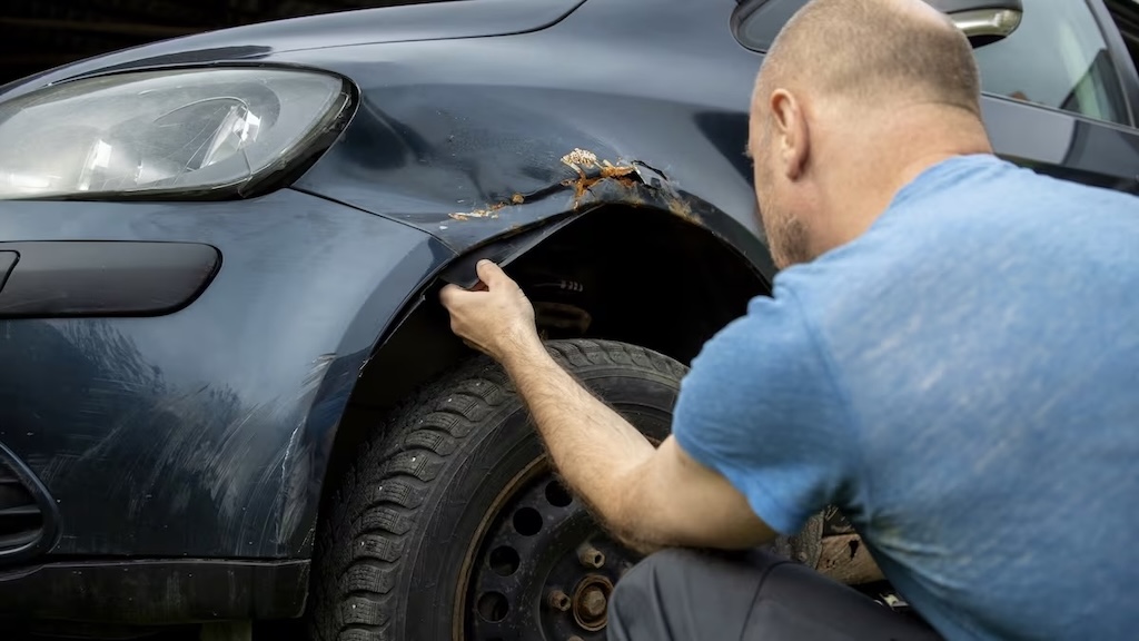 Man inspecting a rusted, damaged front fender on a dark blue car