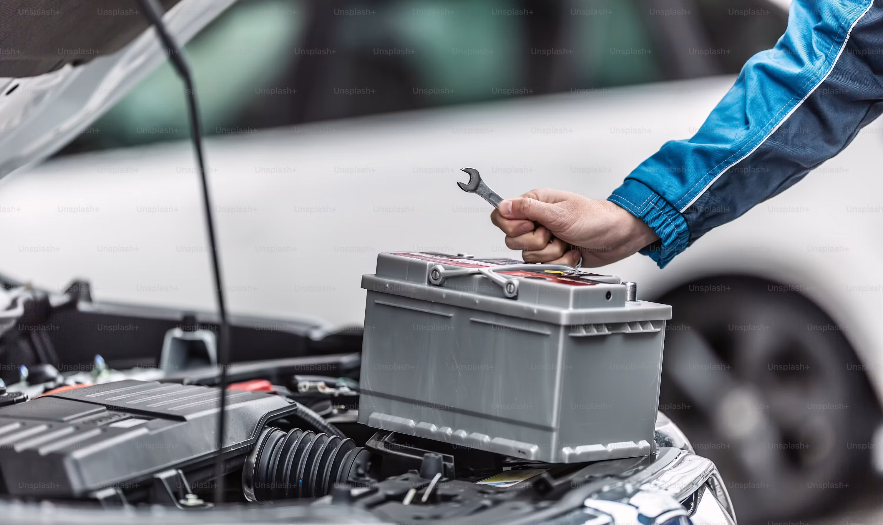 A close-up shot shows a mechanic wearing a blue jacket, holding a small silver wrench in his right hand, poised to work on a grey car battery that is positioned within the open engine bay of a car.