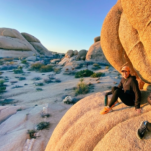 Isabel is sitting on top of large rock in Joshua Tree during sunset. She is smiling at the camera.