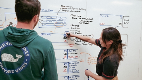 Isabel, on the right, is working a dry erase board with a teammate. She is drawing different possible iterations of an interface.