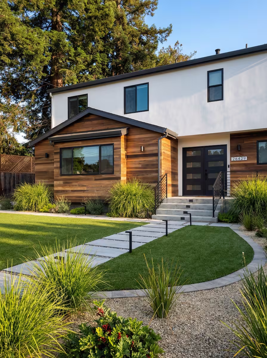 Modern two-story house with white and wooden exterior, a black front door, concrete steps, and a landscaped front yard with green lawn and plants.
