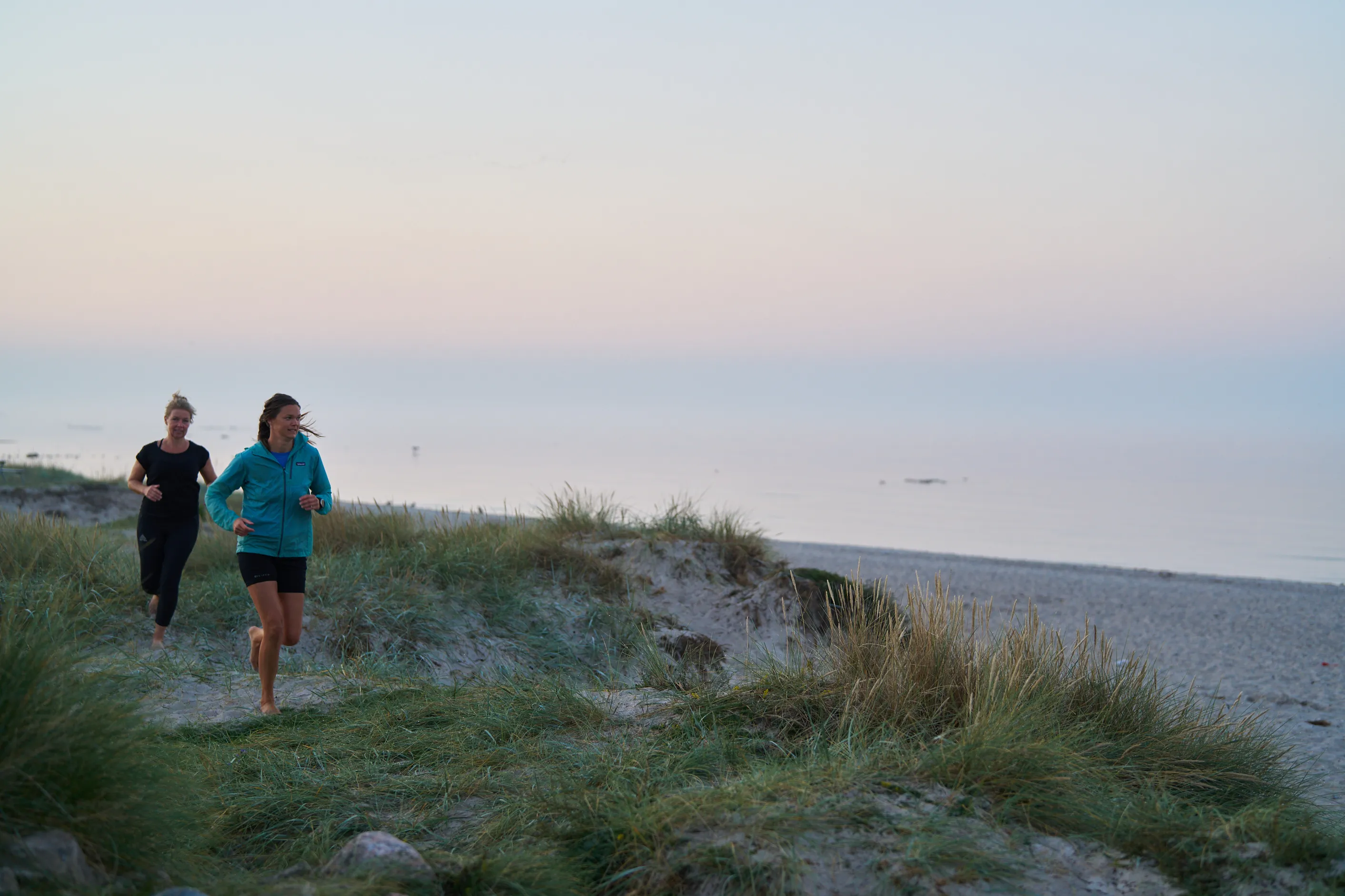 Två löpcoacher springer barfota på en stig genom sanddyner vid en lugn strand under skymningen.