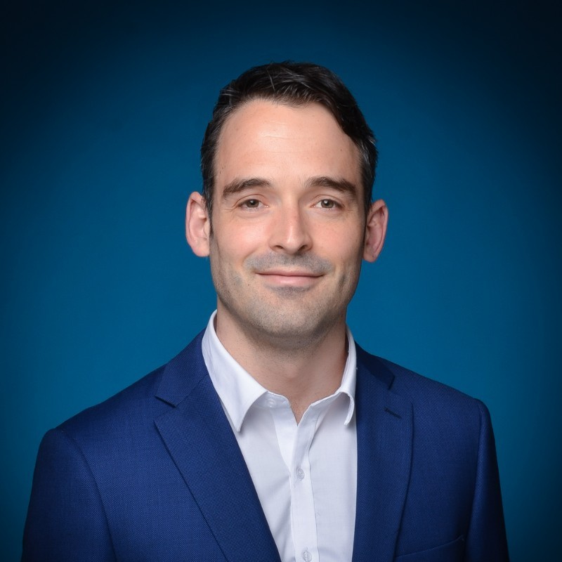 Brendan Hussey wearing a blue blazer and white shirt against a blue background.