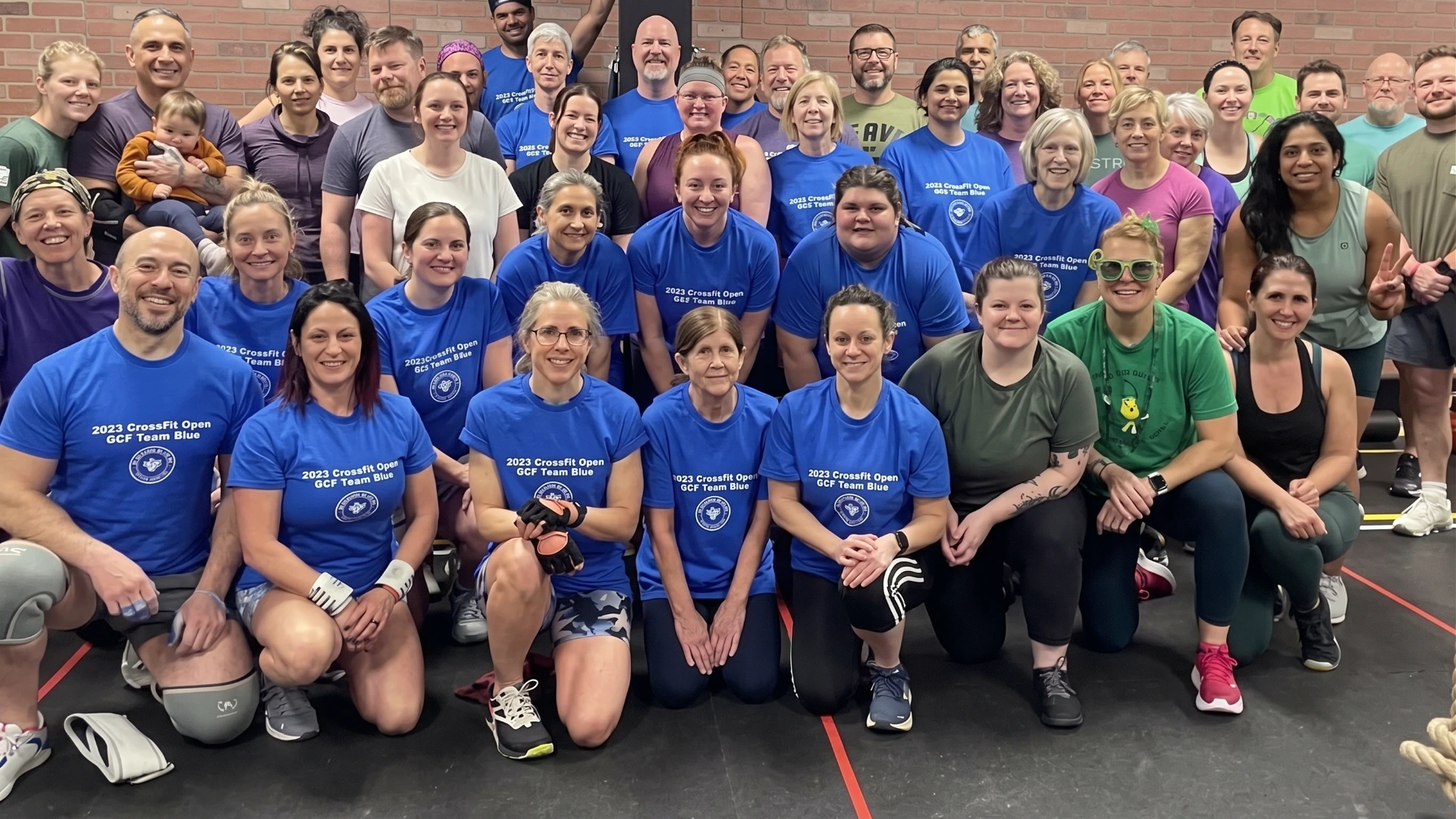 Large group of smiling people posing indoors in front of a brick wall, many wearing matching blue t-shirts that read '2023 CrossFit Open GCF Team Blue,' some kneeling in front while others stand behind, showing diverse ages and genders.