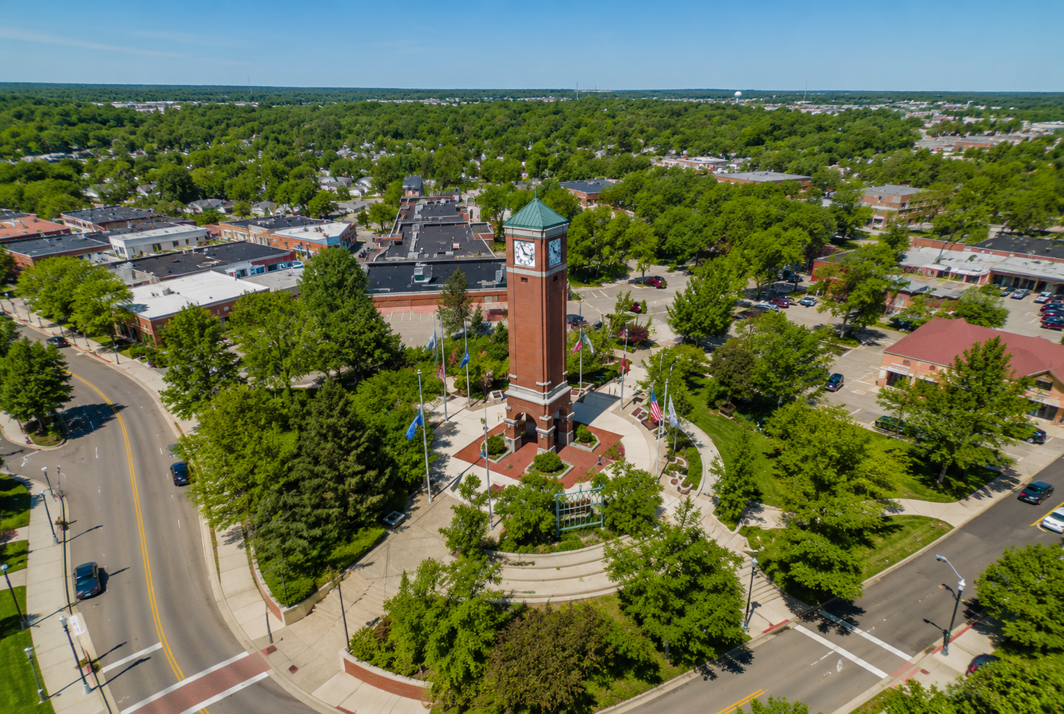 Aerial view of a central brick clock tower surrounded by trees, flags, and roads in a suburban area.
