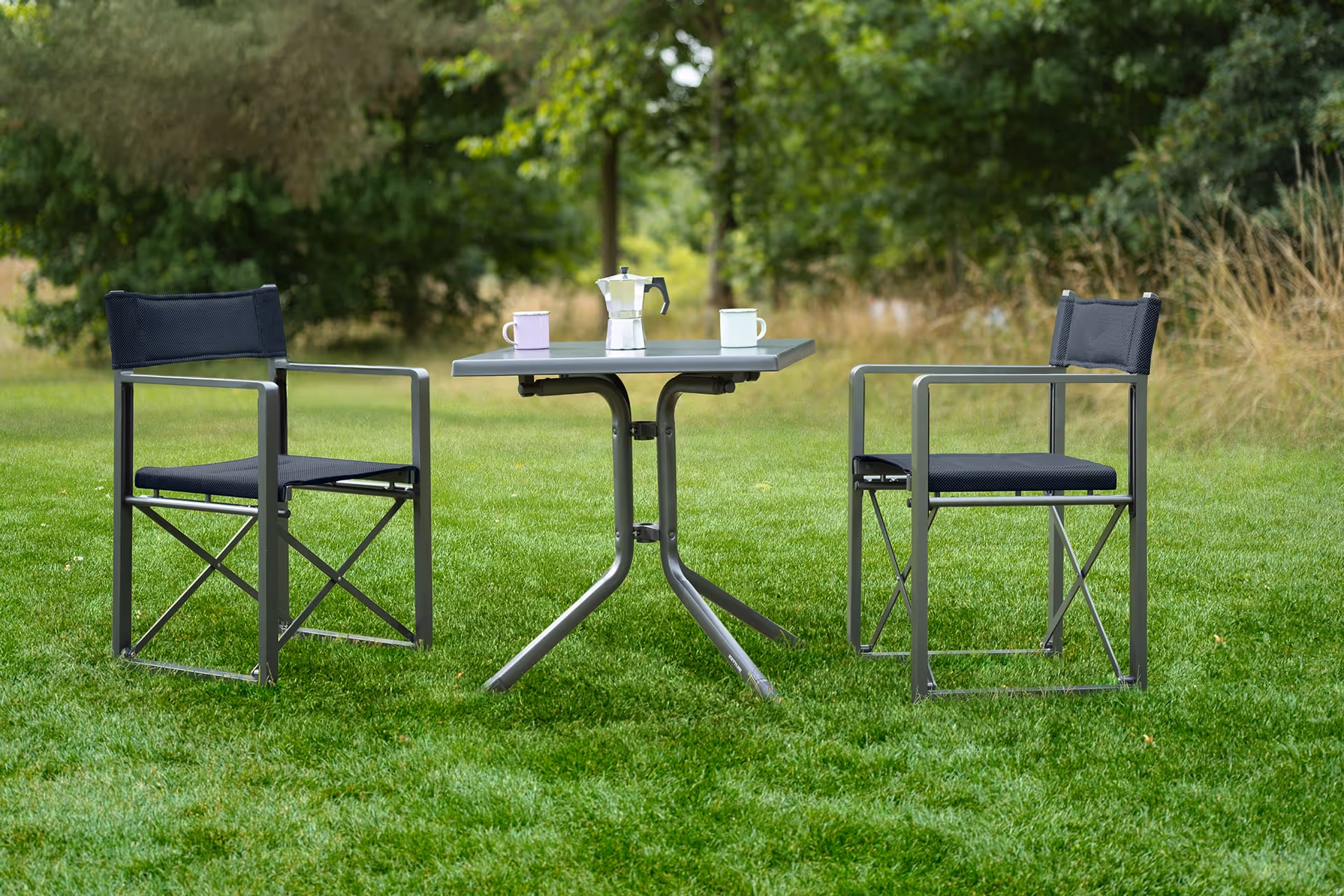 Zwei schwarze NORRLAND Regiestühle und ein kleiner BOULEVARD Tisch mit zwei Tassen und einer Espressokanne auf grünem Gras im Freien.