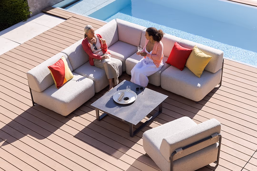Zwei Frauen sitzen auf einer hellen TRIBE Outdoor-Lounge-Eckcouch neben einem Pool und unterhalten sich bei Sonnenschein.