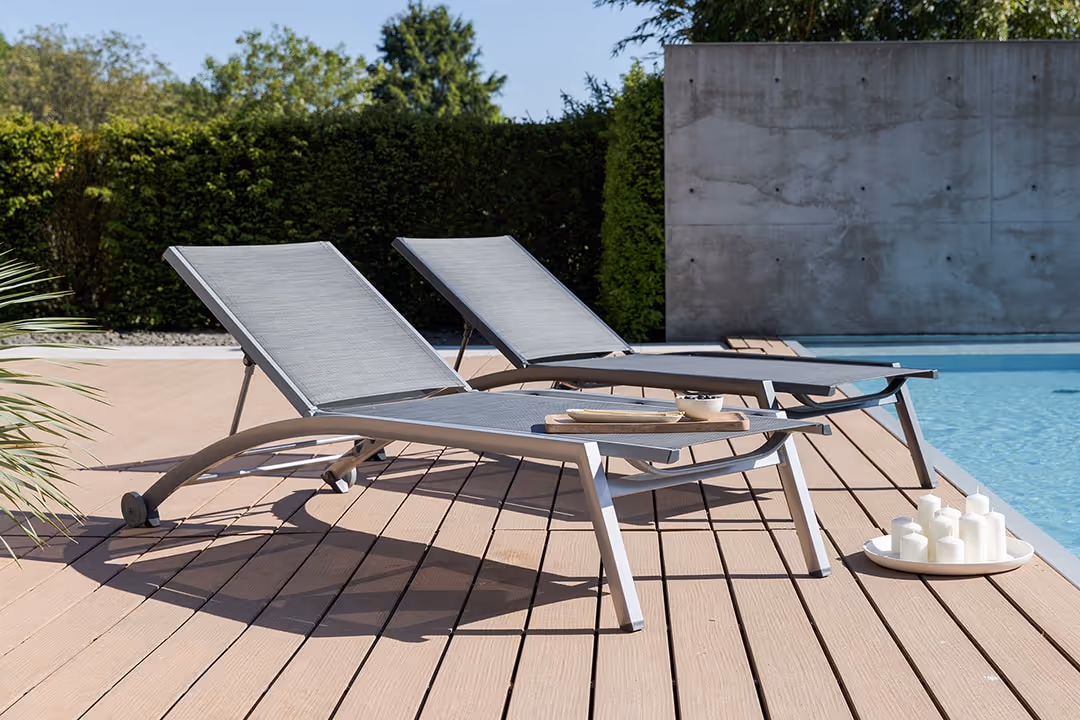 Zwei moderne graue FORMA II Gartenliegen auf Holzterrasse neben Pool mit Kerzentablett.