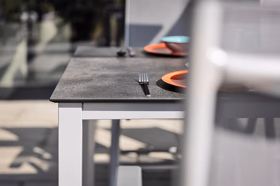 Moderner Esstisch mit dunkler Tischplatte, bestückt mit orangefarbenen Tellern und Besteck, unscharf eine Stuhllehne im Vordergrund.