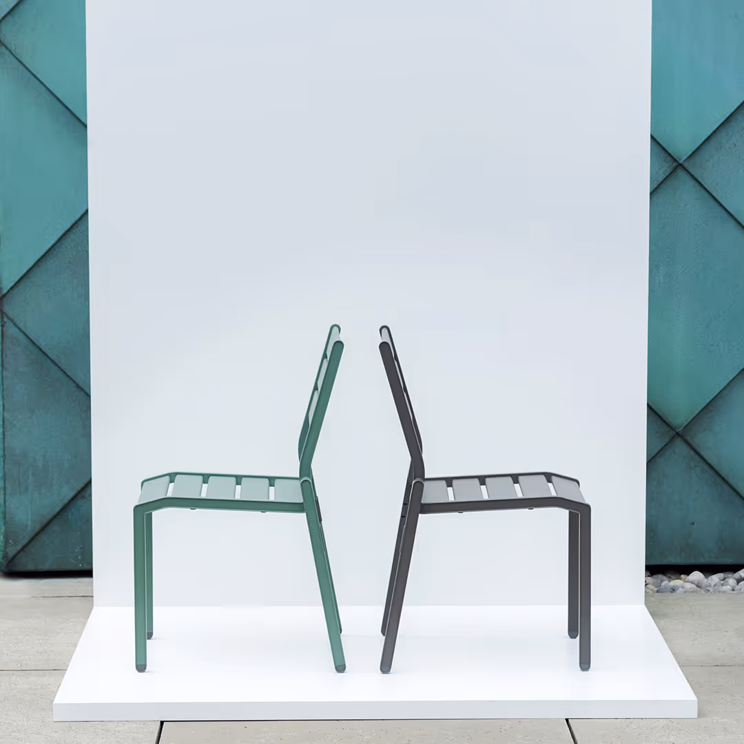 Zwei moderne stapelbare Metallstühle, einer grün und einer schwarz, Rücken an Rücken auf weißer Plattform vor weißer und grüner Wand.