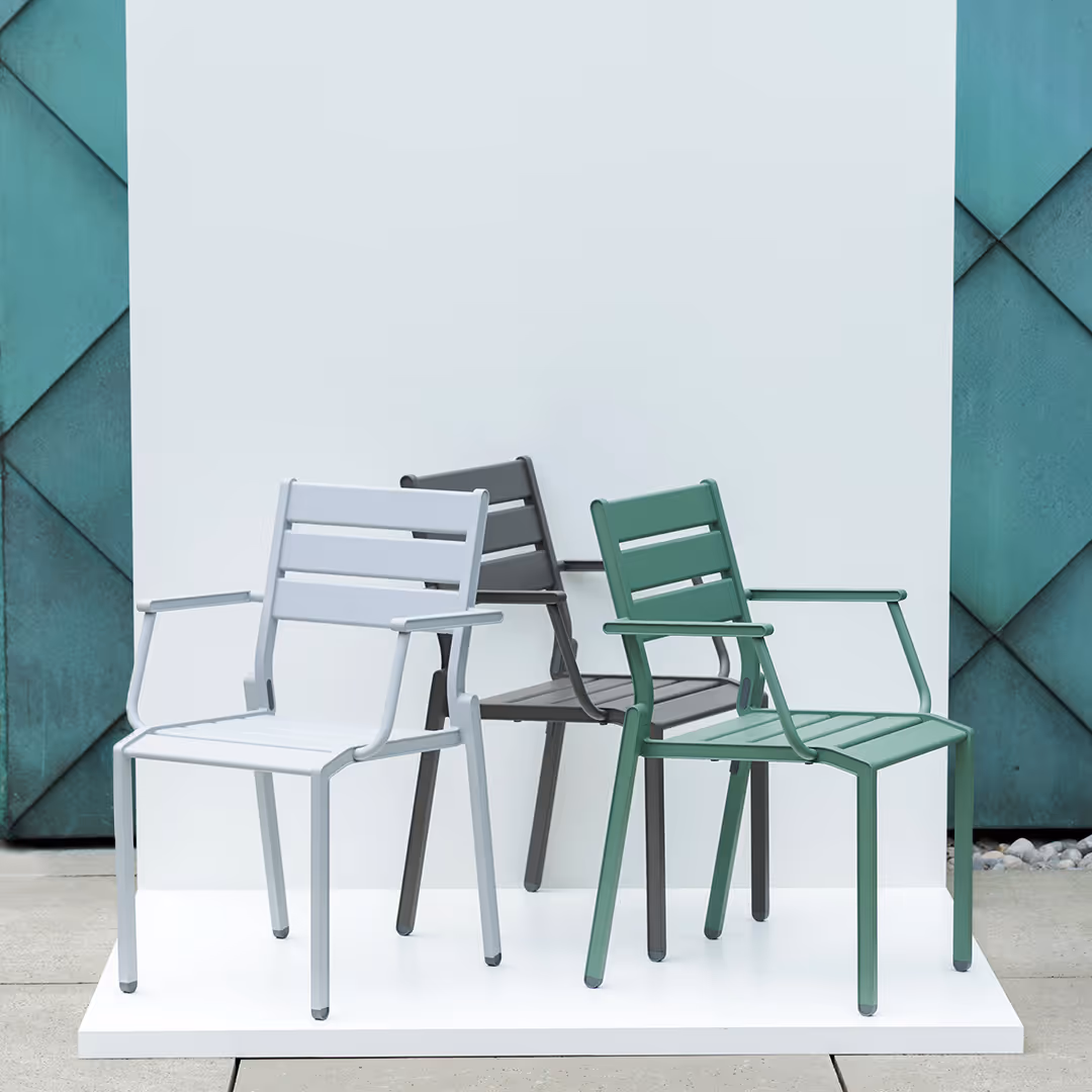 Drei moderne Stapelsessel mit Armlehnen in Grau, Dunkelgrau und Grün auf weißem Podest vor grünlicher Wand.