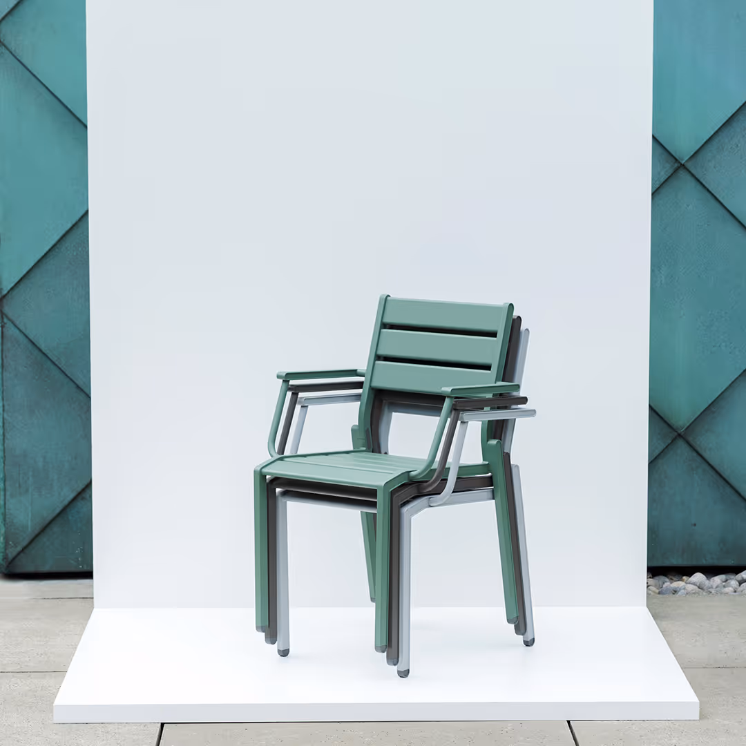 Grüner Gartenstuhl mit Armlehnen, gestapelt vor einem weißen Hintergrund mit grünem, geometrischem Wandmuster.