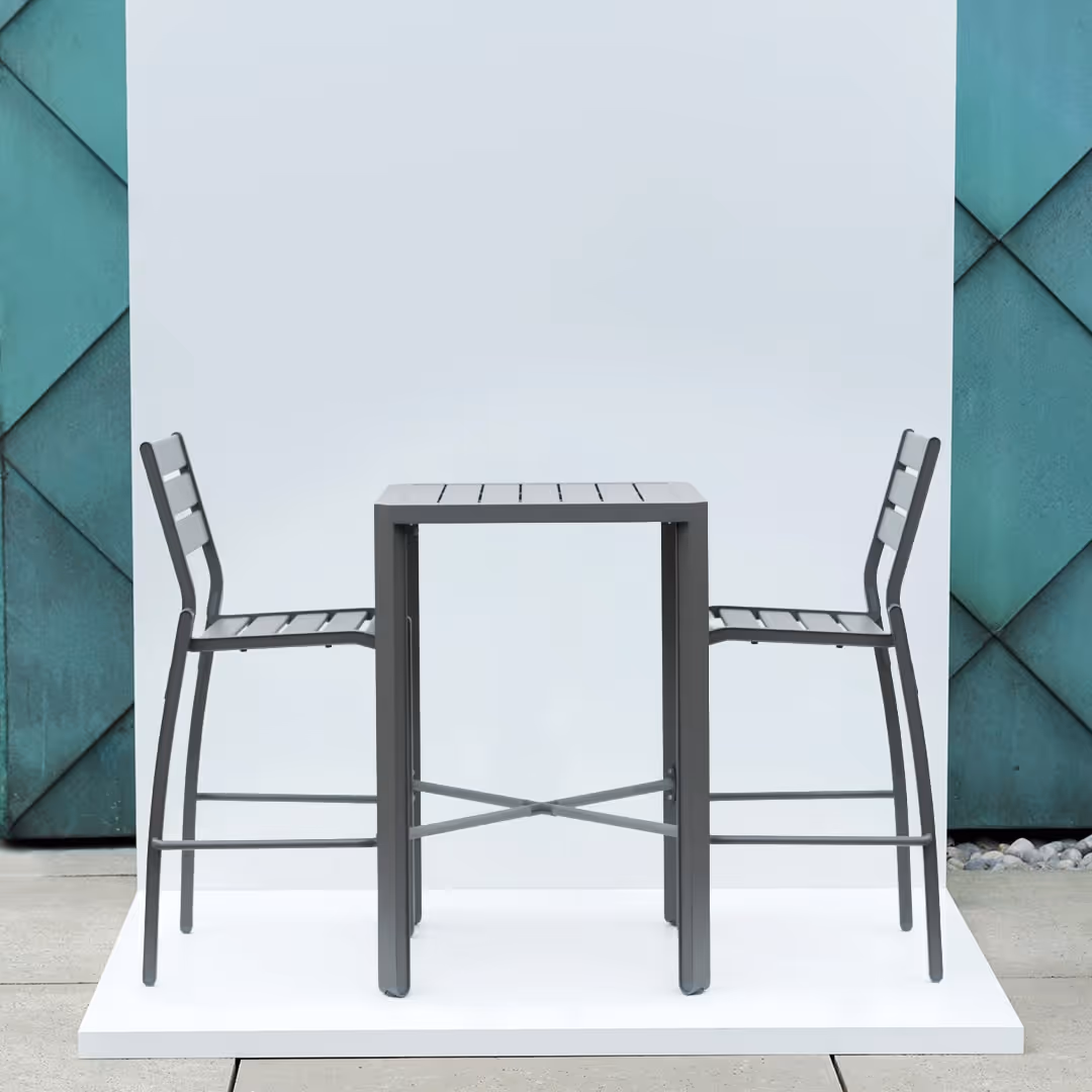 Moderner grauer Bartisch mit zwei passenden Barhockern auf weißer Podestfläche vor grün-blau gefliester Wand.