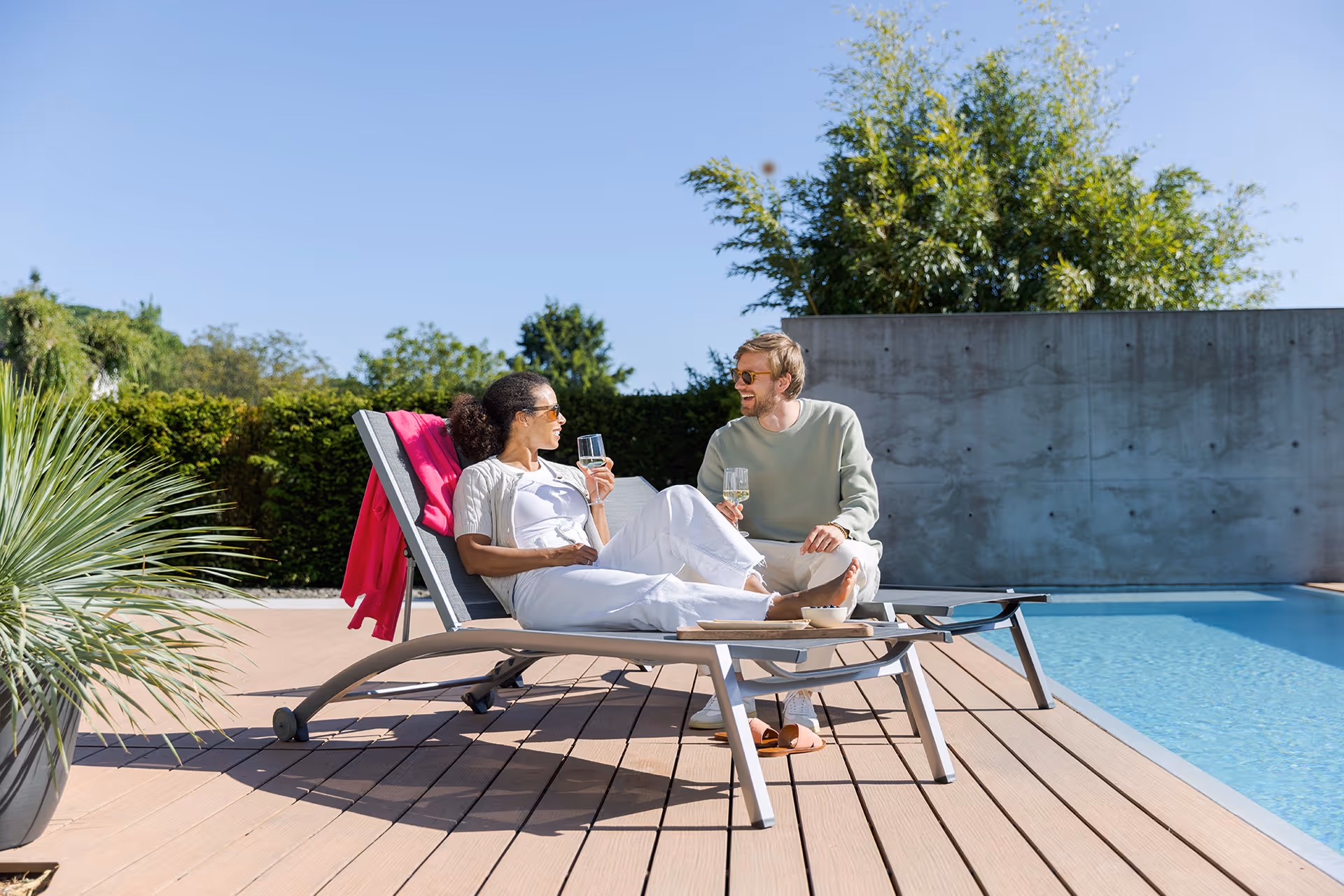 Ein Paar entspannt sich mit Getränken auf Liegestühlen neben einem Pool an einem sonnigen Tag.