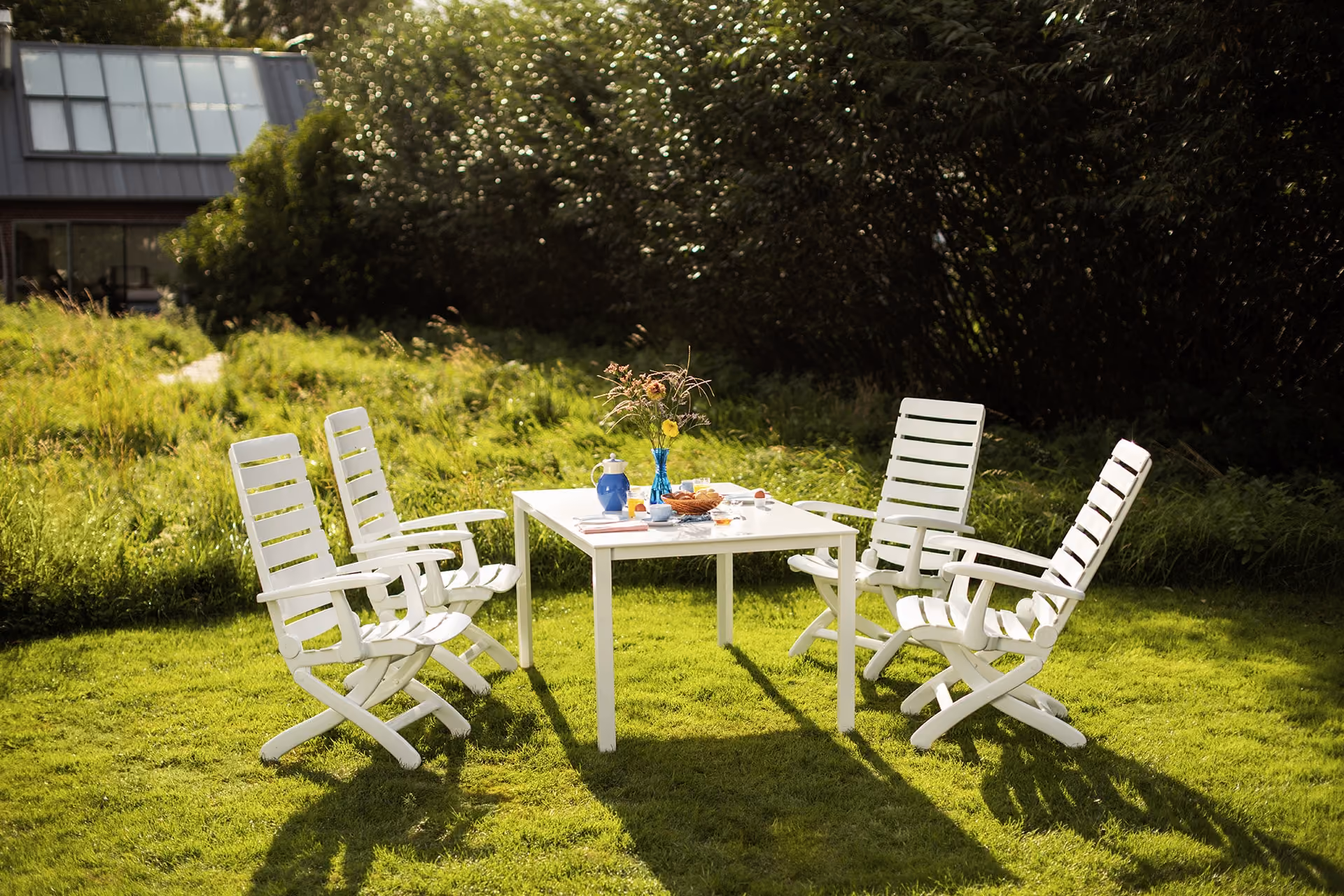 Weiße Gartenmöbel (Tiffany) mit gedecktem Tisch auf einer sonnigen Rasenfläche vor dichter Bepflanzung.