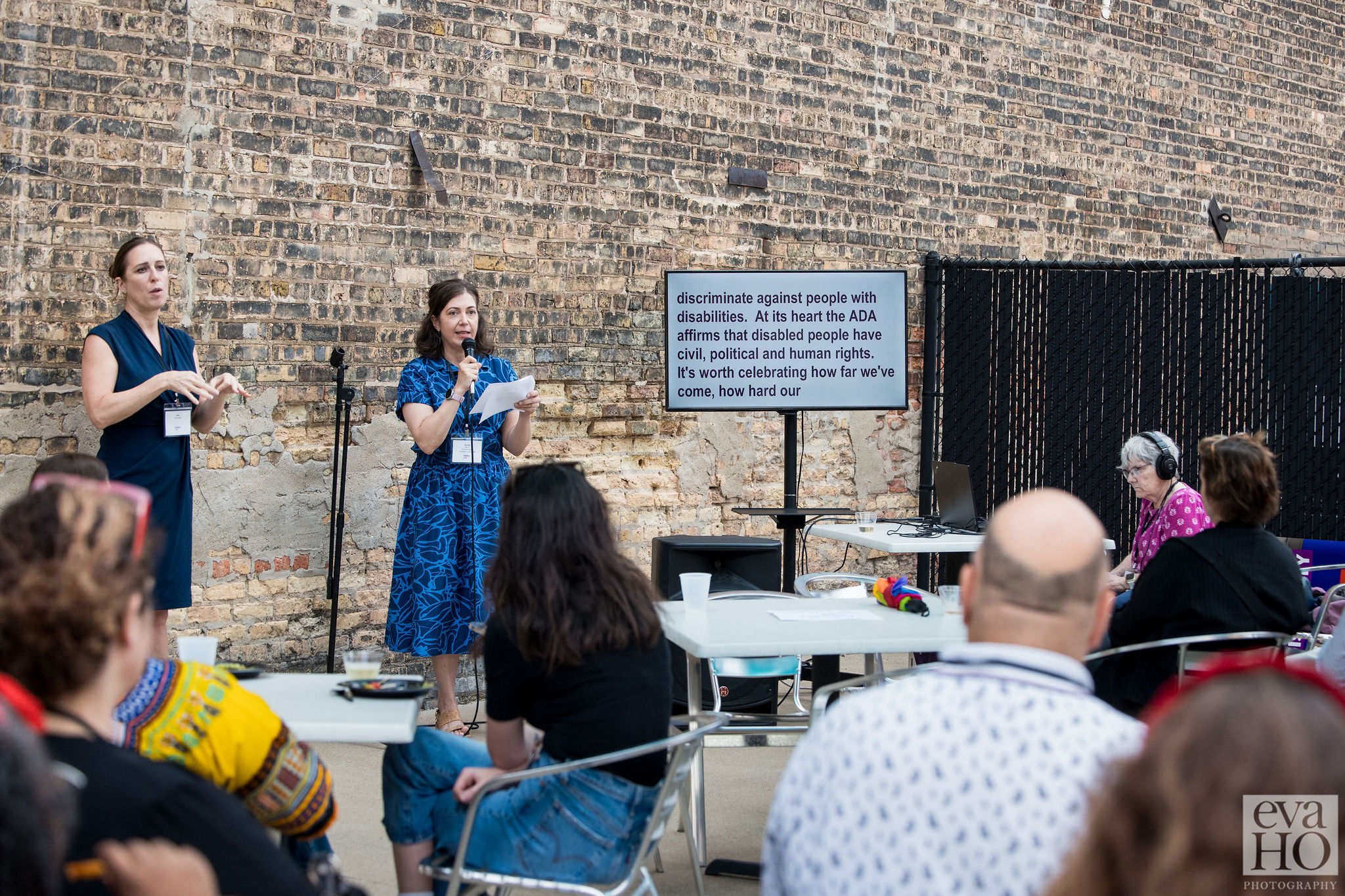 Disability Lead CEO Emily Blum addresses a crowd outdoors in front of a brown brick wall. Emily is wearing a blue dress and is holding a microphone. On her left is a screen with CART and on her right is an ASL interpreter wearing a navy blue dress. The crowd in front of Emily is seated and is of various races, genders, and disabilities.