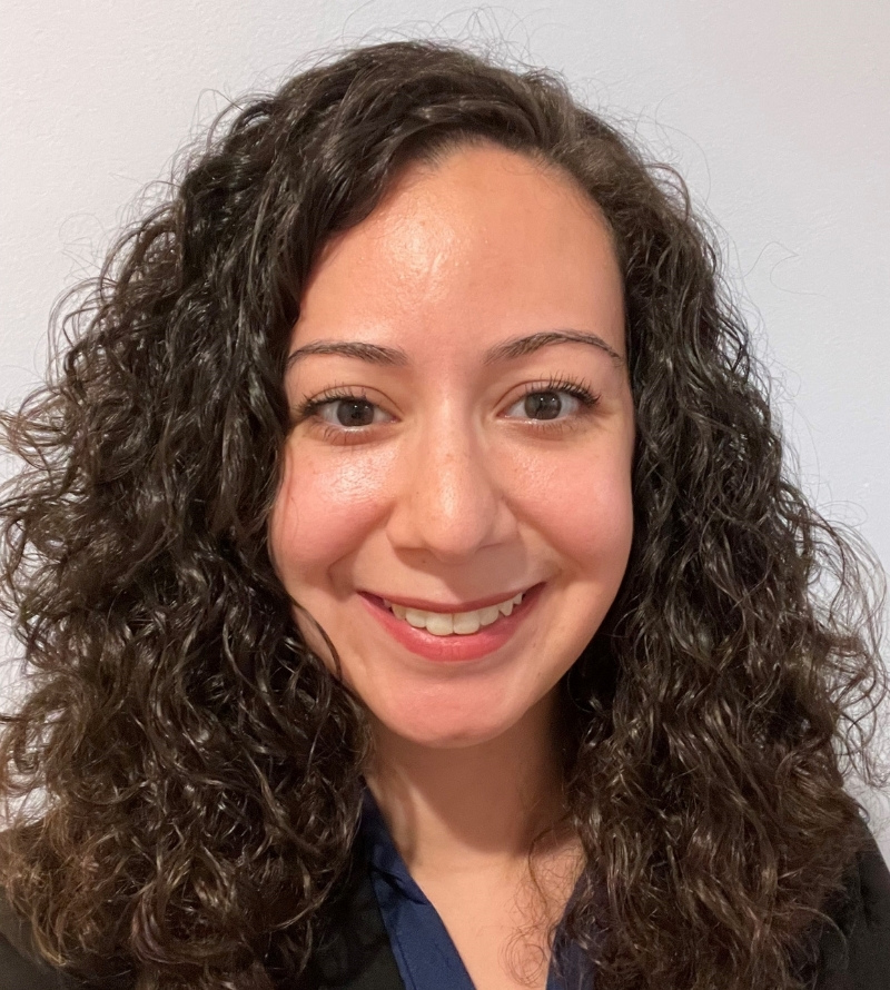  A portrait of Katherine Vakil smiling, a woman with curly brown hair a light complexion wearing a black shirt.